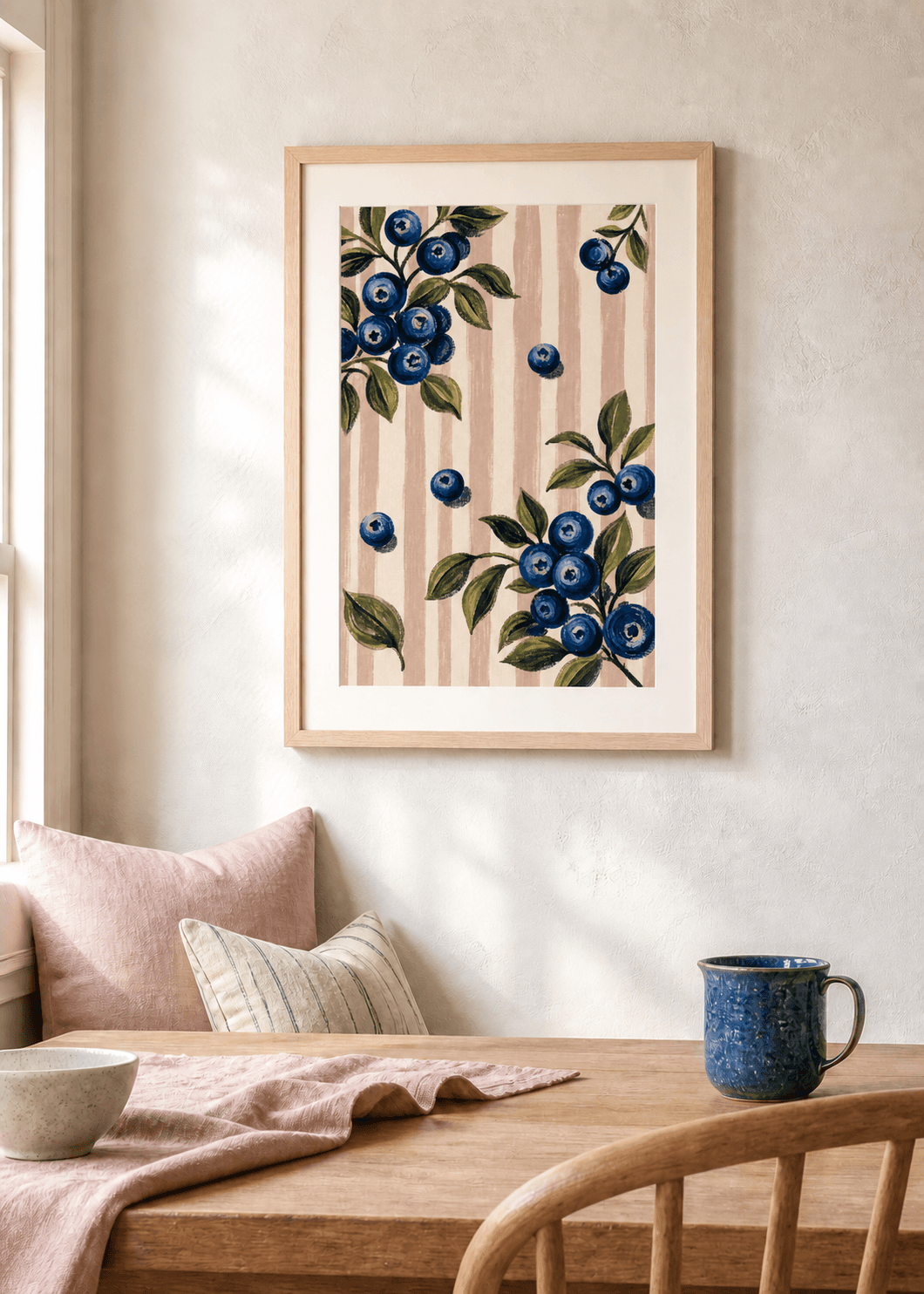 Oak-framed blueberry and blush stripe print above a sunlit breakfast nook with pink linen and a blue stoneware mug